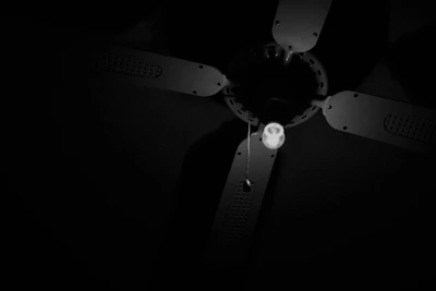 A ceiling fan with four blades and a single illuminated light bulb is suspended from the ceiling. The image is in black and white, with strong contrasts between light and dark areas, highlighting the fan's structure and the pull chain hanging from it.