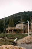 A wooden house with a wraparound balcony is set against a backdrop of a forested mountain. There is a wooden picnic table in the foreground, along with logs and patched grass areas. The house is surrounded by various trees and a serene landscape.
