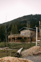 A wooden house with a wraparound balcony is set against a backdrop of a forested mountain. There is a wooden picnic table in the foreground, along with logs and patched grass areas. The house is surrounded by various trees and a serene landscape.