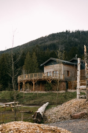 A wooden house with a wraparound balcony is set against a backdrop of a forested mountain. There is a wooden picnic table in the foreground, along with logs and patched grass areas. The house is surrounded by various trees and a serene landscape.