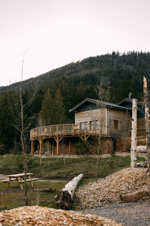 A wooden house with a wraparound balcony is set against a backdrop of a forested mountain. There is a wooden picnic table in the foreground, along with logs and patched grass areas. The house is surrounded by various trees and a serene landscape.