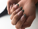 A close-up view of hands displaying elegantly manicured nails with a glossy finish and green marble tips. The skin tone is warm, and the lighting is soft, creating a gentle and sophisticated impression.