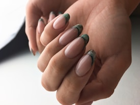 A close-up view of hands displaying elegantly manicured nails with a glossy finish and green marble tips. The skin tone is warm, and the lighting is soft, creating a gentle and sophisticated impression.