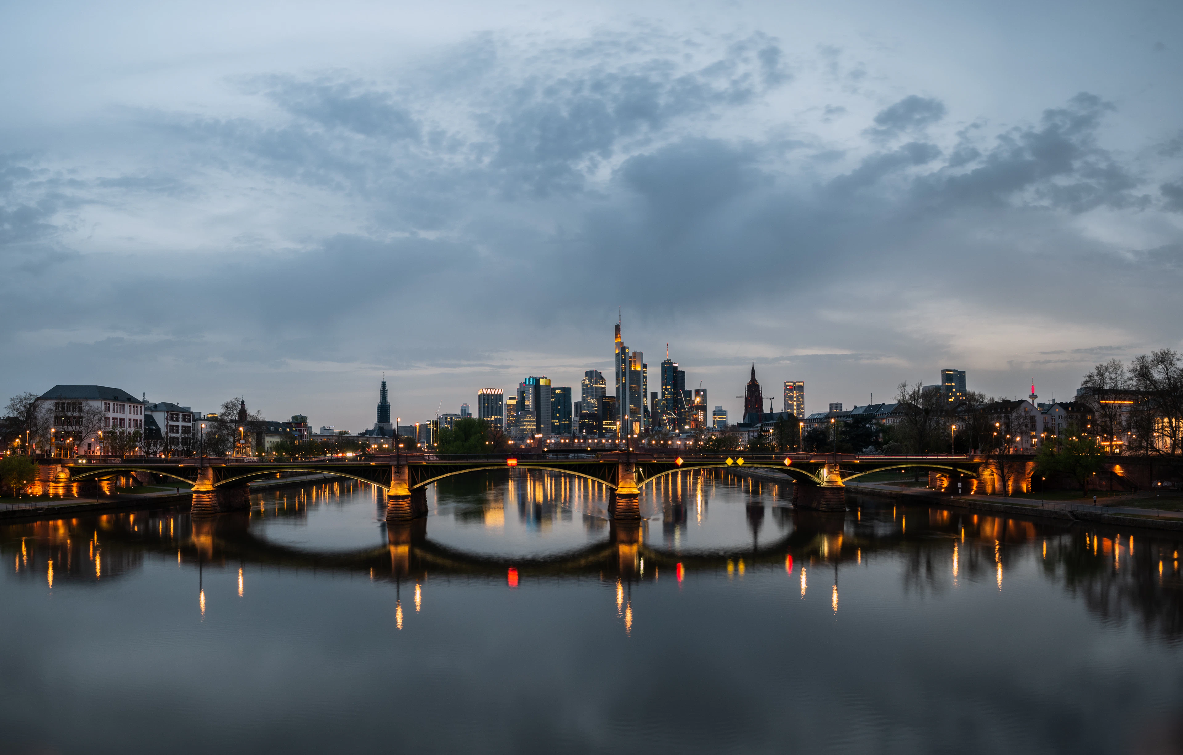 Frankfurt at blue hour seen along the Main, the lit stone arches of the Alte Brücke spanning the river and doubling in the still water, with the banking district's skyline pale behind a heavy grey-blue sky.