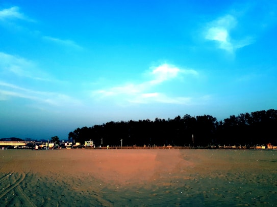 A sandy beach stretches across the foreground with visible tire tracks leading towards a line of trees in the distance. The sky above is a gradient of blue, with scattered clouds adding contrast. A few buildings and structures are seen near the trees, indicating a small occupied area.