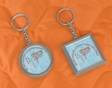 Two keychains lie on an orange quilted fabric background. One keychain is circular, while the other is square. Both have a metallic frame and feature a design with text that reads 'We are exporter of export quality bed sheets' surrounding an image of sheets.