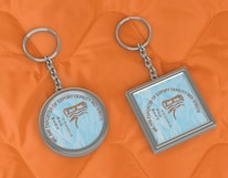 Two keychains lie on an orange quilted fabric background. One keychain is circular, while the other is square. Both have a metallic frame and feature a design with text that reads 'We are exporter of export quality bed sheets' surrounding an image of sheets.