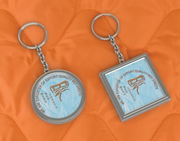 Two keychains lie on an orange quilted fabric background. One keychain is circular, while the other is square. Both have a metallic frame and feature a design with text that reads 'We are exporter of export quality bed sheets' surrounding an image of sheets.