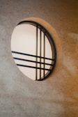 A circular window with a minimalist design features black metal bars crossing over frosted glass, casting shadows on a textured beige wall.
