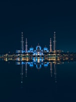 An illuminated mosque with multiple domes and minarets reflected symmetrically on a calm body of water at night. The structure is bathed in blue lighting, creating a serene and majestic atmosphere.