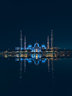 An illuminated mosque with multiple domes and minarets reflected symmetrically on a calm body of water at night. The structure is bathed in blue lighting, creating a serene and majestic atmosphere.