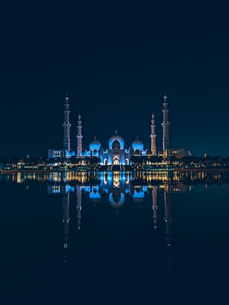 An illuminated mosque with multiple domes and minarets reflected symmetrically on a calm body of water at night. The structure is bathed in blue lighting, creating a serene and majestic atmosphere.