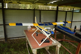 A model airplane with a blue, yellow, and white design sits on a table in a covered outdoor area. The surroundings include corrugated metal sheeting and grass beneath the table. Various equipment and a bottle are placed nearby on the table.