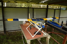 A model airplane with a blue, yellow, and white design sits on a table in a covered outdoor area. The surroundings include corrugated metal sheeting and grass beneath the table. Various equipment and a bottle are placed nearby on the table.