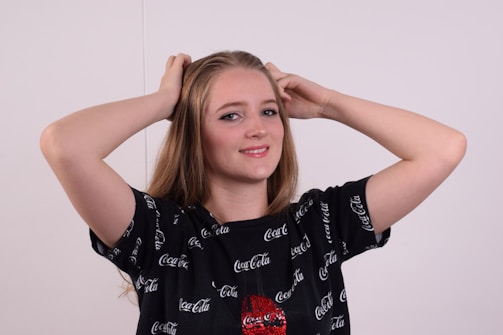 A young woman with long light brown hair smiles while posing with her hands on her head. She is wearing a black T-shirt with the Coca Cola logo printed all over it.