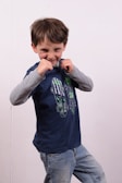 A young child stands in a playful boxing pose, wearing a navy blue t-shirt with skateboard graphics and light blue jeans. The expression is playful with a slight grin, and the background is a plain white wall.