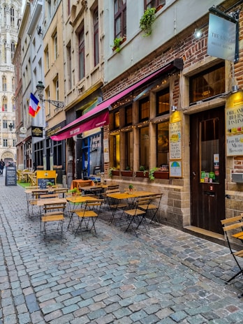 A narrow cobblestone street lined with charming buildings and outdoor cafes. Wooden tables and chairs are arranged on the sidewalk under colorful awnings. The French flag hangs from a building, and a tall, ornate tower is visible in the background. Small plants adorn the window sills.