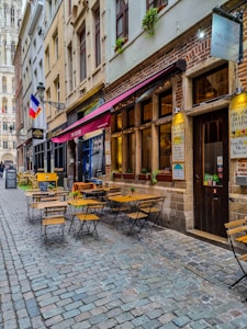 A narrow cobblestone street lined with charming buildings and outdoor cafes. Wooden tables and chairs are arranged on the sidewalk under colorful awnings. The French flag hangs from a building, and a tall, ornate tower is visible in the background. Small plants adorn the window sills.