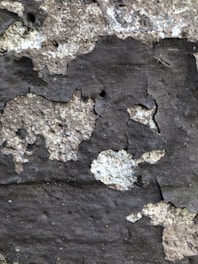 Peeling paint and rough texture of an old wall with uneven surface and patches of darker and lighter shades.