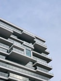 A modern high-rise building with multiple balconies featuring glass railings. The architecture displays a clean, geometric design with a noticeable emphasis on horizontal lines and spacious balconies. The sky is clear with a slightly bluish hue.