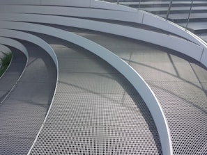 Curved metal grates arranged in a series of parallel, concentric arcs. The surface is sleek and modern, with shadows casting lines across the walkway from a railing. The design is likely part of an architectural structure, showcasing a clean and contemporary aesthetic.