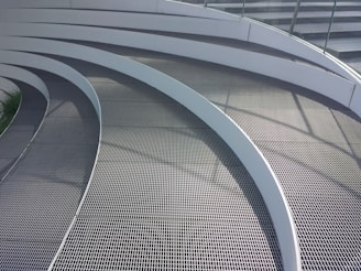 Curved metal grates arranged in a series of parallel, concentric arcs. The surface is sleek and modern, with shadows casting lines across the walkway from a railing. The design is likely part of an architectural structure, showcasing a clean and contemporary aesthetic.