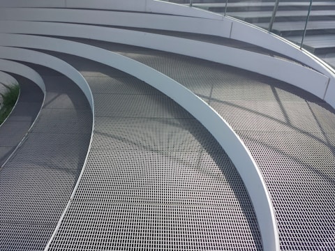 Curved metal grates arranged in a series of parallel, concentric arcs. The surface is sleek and modern, with shadows casting lines across the walkway from a railing. The design is likely part of an architectural structure, showcasing a clean and contemporary aesthetic.