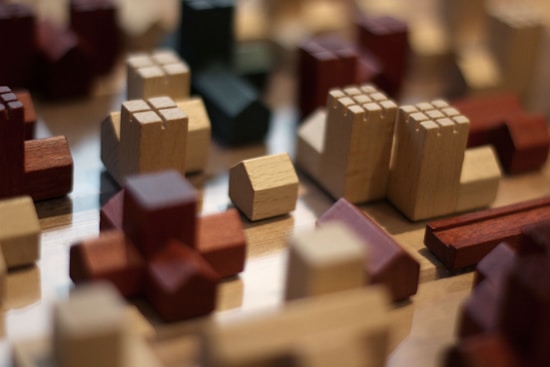 A collection of small wooden blocks in various shapes and colors is arranged on a smooth surface. The shapes resemble buildings, with a juxtaposition of reds, creams, and greens. The focus is on the foreground, with several details such as engraved lines representing windows. The background is blurred, creating a depth of field effect.