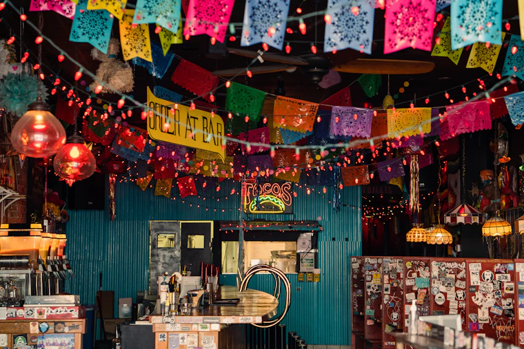 A restaurant bar strung with papel picado banners and warm string lights above a teal wood counter