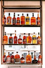 A collection of various whiskey bottles arranged on a three-tier shelf. The bottles come in different shapes, sizes, and label designs, showcasing a range of whiskey brands. Sunlight filters through the window in the background, casting a warm glow on the bottles, enhancing their amber hues.