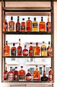 A collection of various whiskey bottles arranged on a three-tier shelf. The bottles come in different shapes, sizes, and label designs, showcasing a range of whiskey brands. Sunlight filters through the window in the background, casting a warm glow on the bottles, enhancing their amber hues.