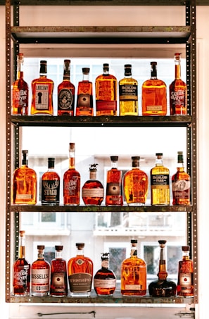 A collection of various whiskey bottles arranged on a three-tier shelf. The bottles come in different shapes, sizes, and label designs, showcasing a range of whiskey brands. Sunlight filters through the window in the background, casting a warm glow on the bottles, enhancing their amber hues.