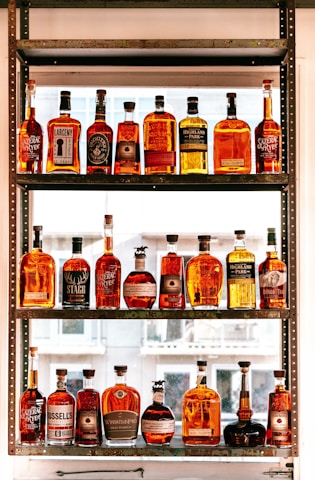 A collection of various whiskey bottles arranged on a three-tier shelf. The bottles come in different shapes, sizes, and label designs, showcasing a range of whiskey brands. Sunlight filters through the window in the background, casting a warm glow on the bottles, enhancing their amber hues.