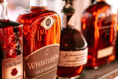 A row of whiskey bottles lined up closely together, showcasing various brands and styles. The bottles have a warm amber color, and some labels are visible, indicating the whiskey's age and origin.