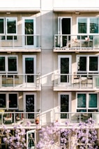 The image depicts a modern apartment building facade with multiple balconies. Each balcony is furnished with chairs and small tables. Below, blooming purple flowers partially obscure the view, adding a touch of nature to the urban setting.