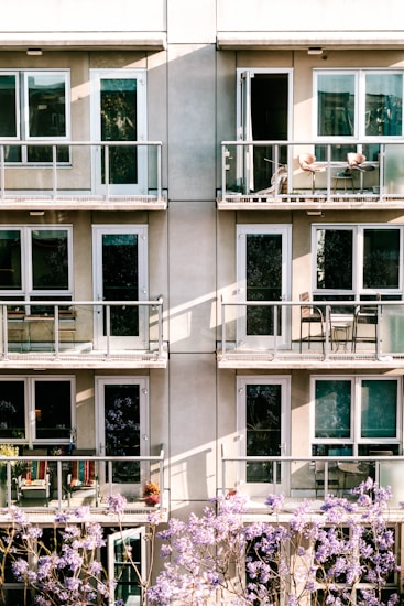 The image depicts a modern apartment building facade with multiple balconies. Each balcony is furnished with chairs and small tables. Below, blooming purple flowers partially obscure the view, adding a touch of nature to the urban setting.