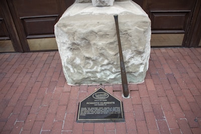A bronze plaque and baseball bat are displayed on a brick sidewalk. The plaque is dedicated to Roberto Clemente and includes details about his baseball achievements. The bat leans against a large stone pillar in front of a wooden facade.