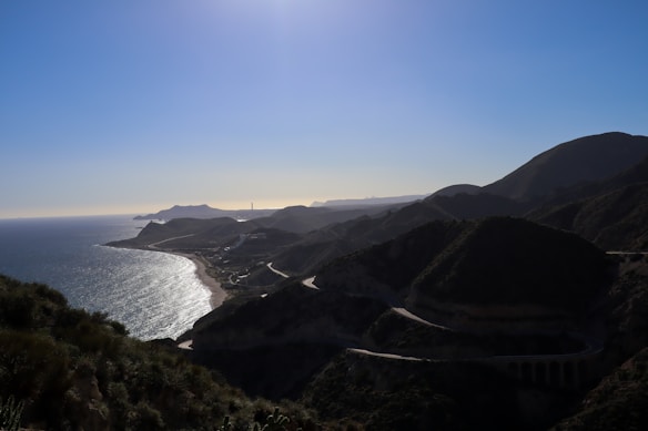 A coastal landscape with a winding road running along hillsides, overlooking a sea that stretches to the horizon. The sunlight casts shadows on the rolling hills and the ocean glimmers under the bright sky.