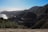 A winding coastal road snakes through rugged, mountainous terrain. The road curves around the hillsides, and in the foreground, there are dense green cacti. The sea is visible to the left, with waves gently lapping against the shore. The distant horizon reveals layers of hills fading into the soft light of the sky.
