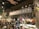A cozy coffee shop interior featuring a large sign that reads 'Coffee Project'. The setting includes a counter with a display of various pastries and colorful flower arrangements. Wooden and metal elements are prominent in the architecture, along with warm lighting and decorative foliage.
