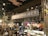 A cozy coffee shop interior featuring a large sign that reads 'Coffee Project'. The setting includes a counter with a display of various pastries and colorful flower arrangements. Wooden and metal elements are prominent in the architecture, along with warm lighting and decorative foliage.