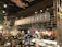 A cozy coffee shop interior featuring a large sign that reads 'Coffee Project'. The setting includes a counter with a display of various pastries and colorful flower arrangements. Wooden and metal elements are prominent in the architecture, along with warm lighting and decorative foliage.