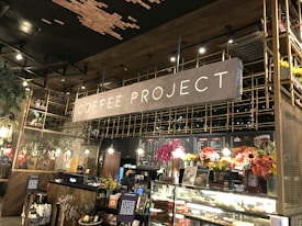 A cozy coffee shop interior featuring a large sign that reads 'Coffee Project'. The setting includes a counter with a display of various pastries and colorful flower arrangements. Wooden and metal elements are prominent in the architecture, along with warm lighting and decorative foliage.