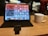 A tablet on a stand displays various app icons and widgets on its screen. Next to it is a white coffee mug on a wooden table. The background is blurred, showing soft lights and part of a person.