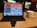 A tablet on a stand displays various app icons and widgets on its screen. Next to it is a white coffee mug on a wooden table. The background is blurred, showing soft lights and part of a person.