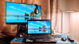 A dual-monitor setup is on a desk with a larger monitor placed behind a laptop. Both screens display the same wallpaper of a beach with a rock formation and a person running. There is a pair of headphones resting on the corner of the monitor. A desk lamp is positioned to the right, and a mouse with a pad is beside the laptop.