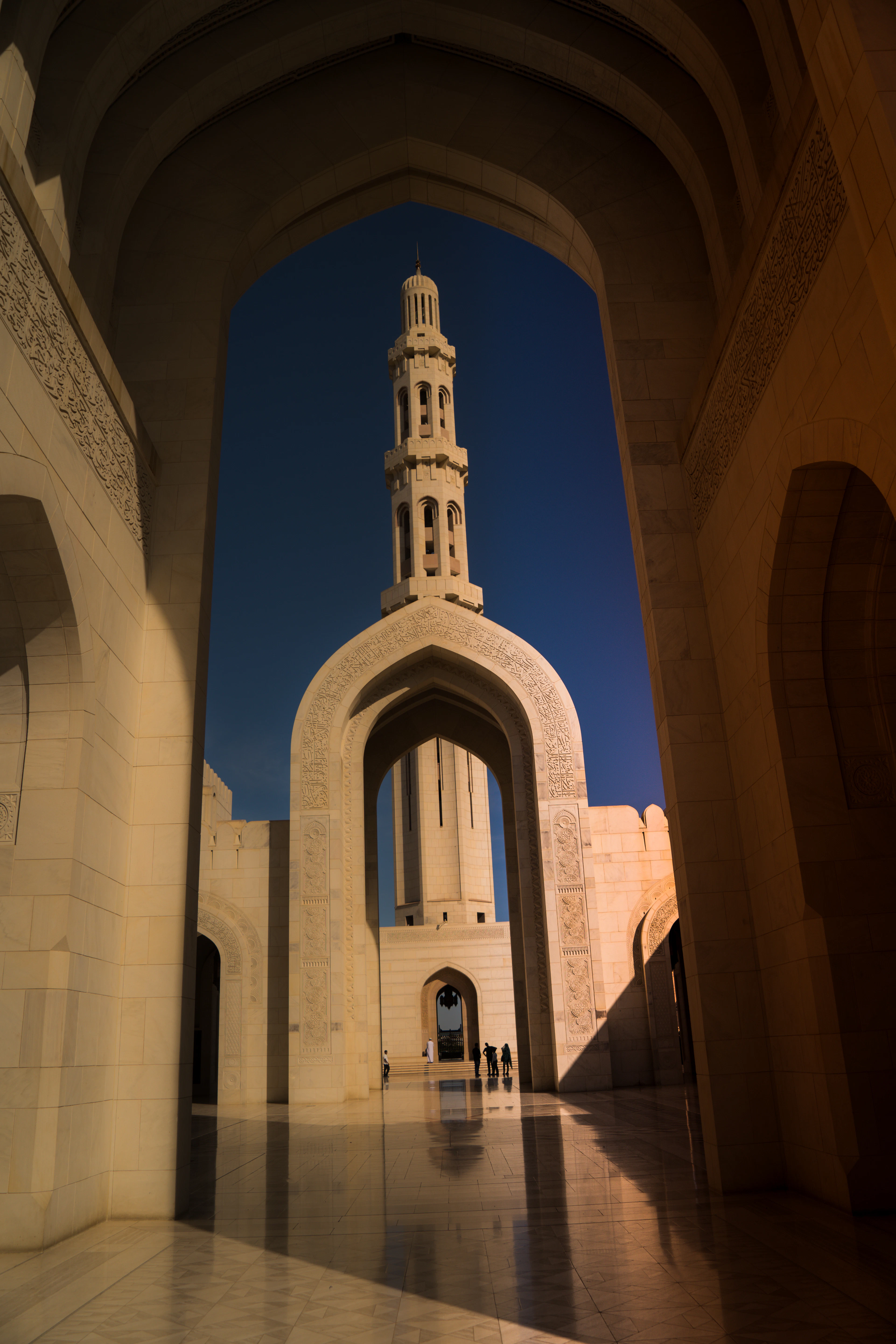An archway inside a mosque frames a tall minaret against a deep blue sky, raking light on the stone.