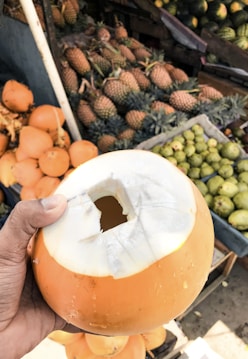 A person's hand is holding a partially peeled yellow coconut with a square opening on the top. In the background, there are crates filled with various fruits, including pineapples, limes, and more yellow coconuts.
