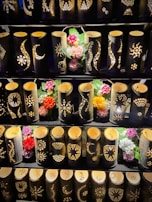 A display of bamboo lanterns with intricate patterns carved into their surfaces, illuminated from within. The lanterns are arranged in rows and accompanied by colorful artificial flowers interspersed among them.