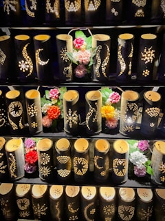 A display of bamboo lanterns with intricate patterns carved into their surfaces, illuminated from within. The lanterns are arranged in rows and accompanied by colorful artificial flowers interspersed among them.